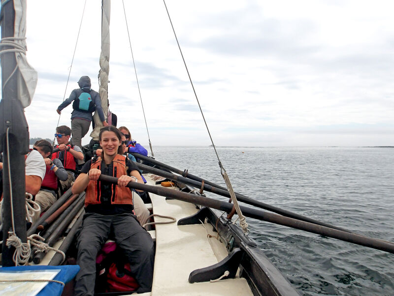 A group of people are on a boat, possibly a sailboat, on the water. A woman in the foreground is wearing a life jacket and holding onto a long oar or pole. Other people are visible on the boat, and the sea stretches out to the horizon under a cloudy sky. The boat appears to be in motion, and the overall scene suggests a maritime adventure or excursion.